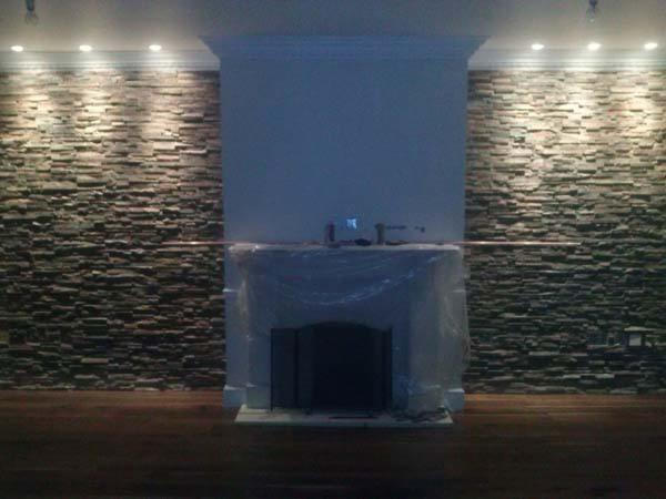 Ściana z kamienia dekoracyjnego z podświetleniem LED, osłonięty kominek, drewniana podłoga. Etap prac wykończeniowych, folia ochronna na kominku.