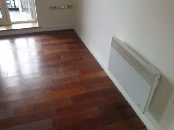 Błyszczący, ciemny parkiet w pokoju z białymi ścianami i grzejnikiem elektrycznym. Okno w lewym górnym rogu odbija światło na podłodze.