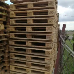 Stos drewnianych palet EPAL ułożonych jeden na drugim, widoczny fragment ogrodzenia z siatki i zieleń w tle.