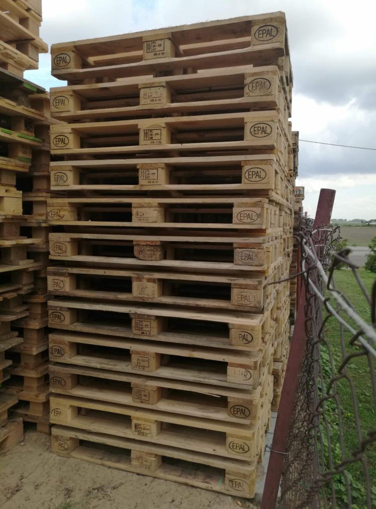 Stos drewnianych palet EPAL ułożonych jeden na drugim, widoczny fragment ogrodzenia z siatki i zieleń w tle.