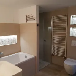 Jasna łazienka z beżowymi płytkami, mozaiką w podświetlonych wnękach, grzejnikiem drabinkowym i kabiną prysznicową z kamienną posadzką.