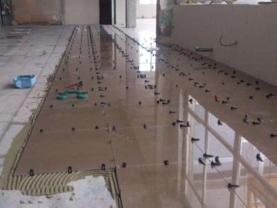 Układanie brązowych płytek podłogowych z użyciem klipsów poziomujących. Widoczne narzędzia i klej. Praca w trakcie realizacji, jasne wnętrze.