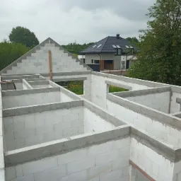 Wznoszenie ścian z białych bloczków betonu komórkowego, widoczne zbrojenie nadproży, w tle dom jednorodzinny pod czarnym dachem i zieleń.