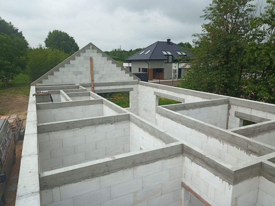 Wznoszenie ścian z białych bloczków betonu komórkowego, widoczne zbrojenie nadproży, w tle dom jednorodzinny pod czarnym dachem i zieleń.