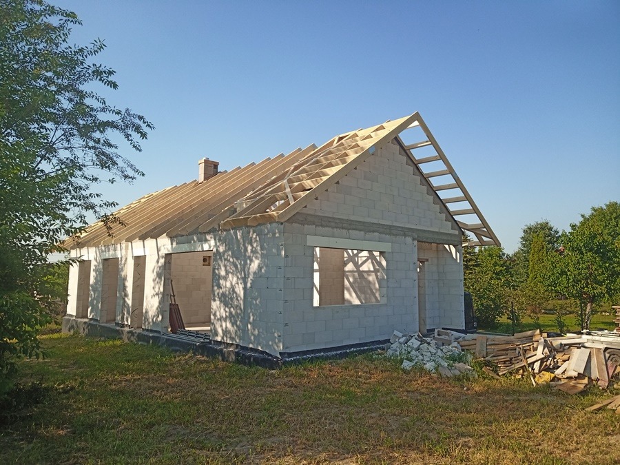 Surowy dom murowany z bloczków z widoczną konstrukcją dachu, ceglany komin i porozrzucane materiały budowlane na trawie.