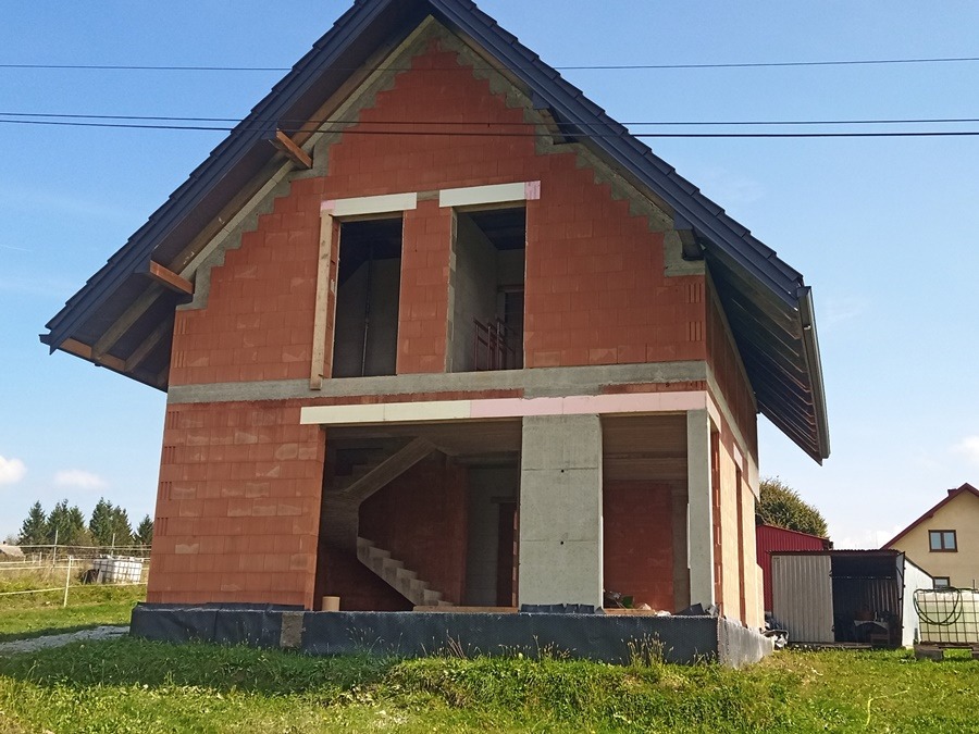 Surowy stan domu z czerwonej cegły z widocznymi schodami wewnętrznymi, zabezpieczony czarną folią fundamentową, z częściowo zamontowanym dachem i oknami bez szyb.