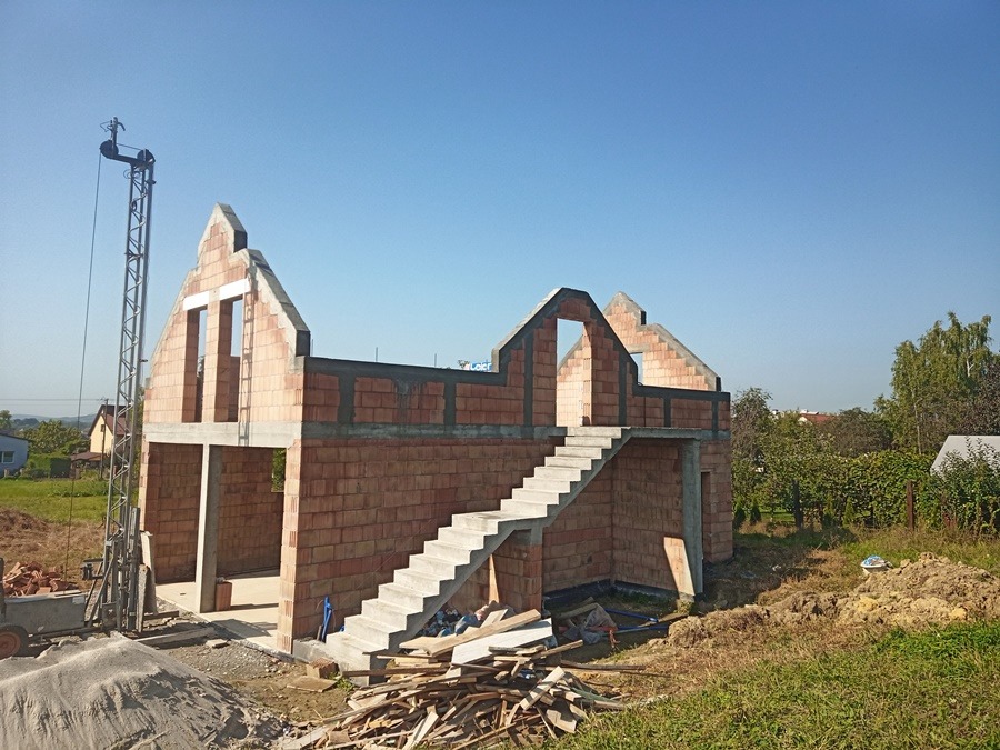 Surowy stan domu z czerwonej cegły z widocznymi schodami zewnętrznymi, dźwigiem i stertą drewna na pierwszym planie w słoneczny dzień.