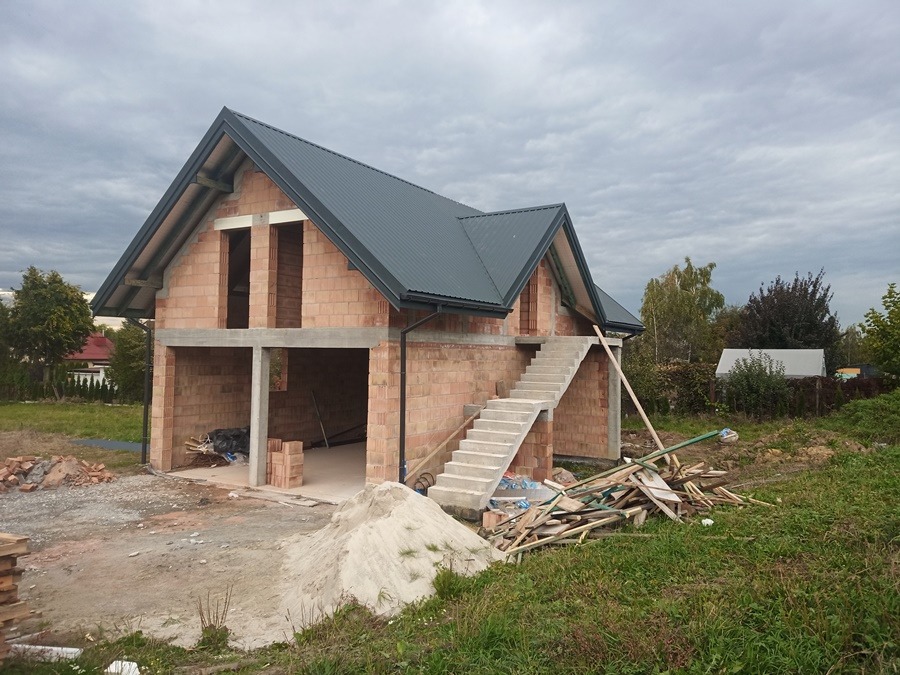 Surowy stan budynku z czerwonej cegły z szarym dachem, widoczne betonowe schody wejściowe, stos cegieł i sterta drewna na trawniku, pochmurne niebo.