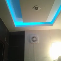 Nowoczesny sufit podwieszany z niebieskim oświetleniem LED w łazience. Widoczny wentylator na ścianie z białych płytek i fragment ciemnej zabudowy.