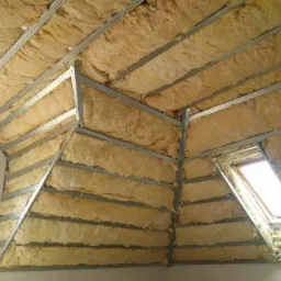 Izolacja poddasza wełną mineralną w metalowym stelażu, widoczne okno dachowe i fragment ściany.