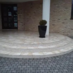 Wyłożone płytkami schody wejściowe z trzema stopniami, wykończone mozaiką, prowadzące do drzwi wejściowych, z kolumną i donicą obok.