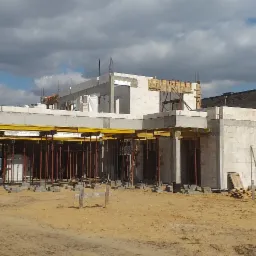 Wznoszenie ścian z bloczków betonowych na budowie domu jednorodzinnego, widoczne deskowanie i podpory stropu, w tle pochmurne niebo i sąsiedni budynek.