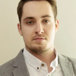 Portret młodego mężczyzny w szarej marynarce i koszuli w kropki, z delikatnym zarostem i niebieskimi oczami, na beżowym tle.