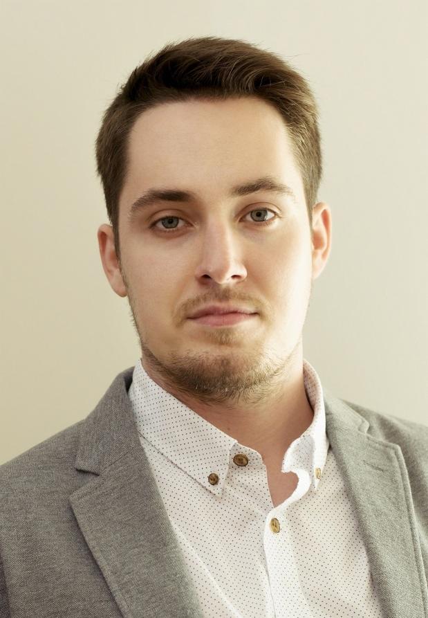 Portret młodego mężczyzny w szarej marynarce i koszuli w kropki, z delikatnym zarostem i niebieskimi oczami, na beżowym tle.