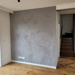 Szara ściana z efektem dekoracyjnym, drewniana podłoga ułożona w jodełkę, widoczne schody w tle, gniazdka elektryczne przy listwie przypodłogowej.