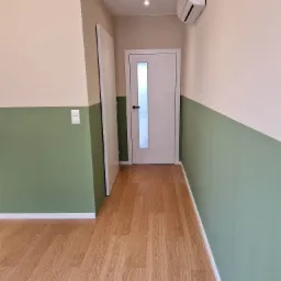 Wnętrze pokoju po remoncie z podziałem kolorystycznym ścian: g&oacute;ra beżowa, d&oacute;ł zielony, podłoga z paneli imitujących drewno, białe drzwi z przeszkleniem i klimatyzator na ścianie.