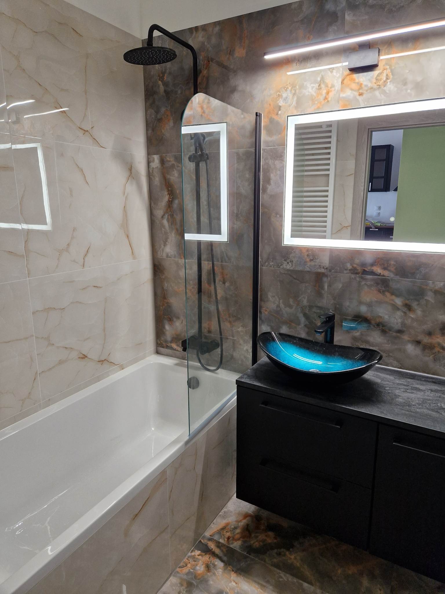 Nowoczesna łazienka z marmurowymi płytkami, czarną armaturą, podświetlanym lustrem i umywalką w odcieniach niebieskiego, widok na kabinę prysznicową za szklaną ścianką.