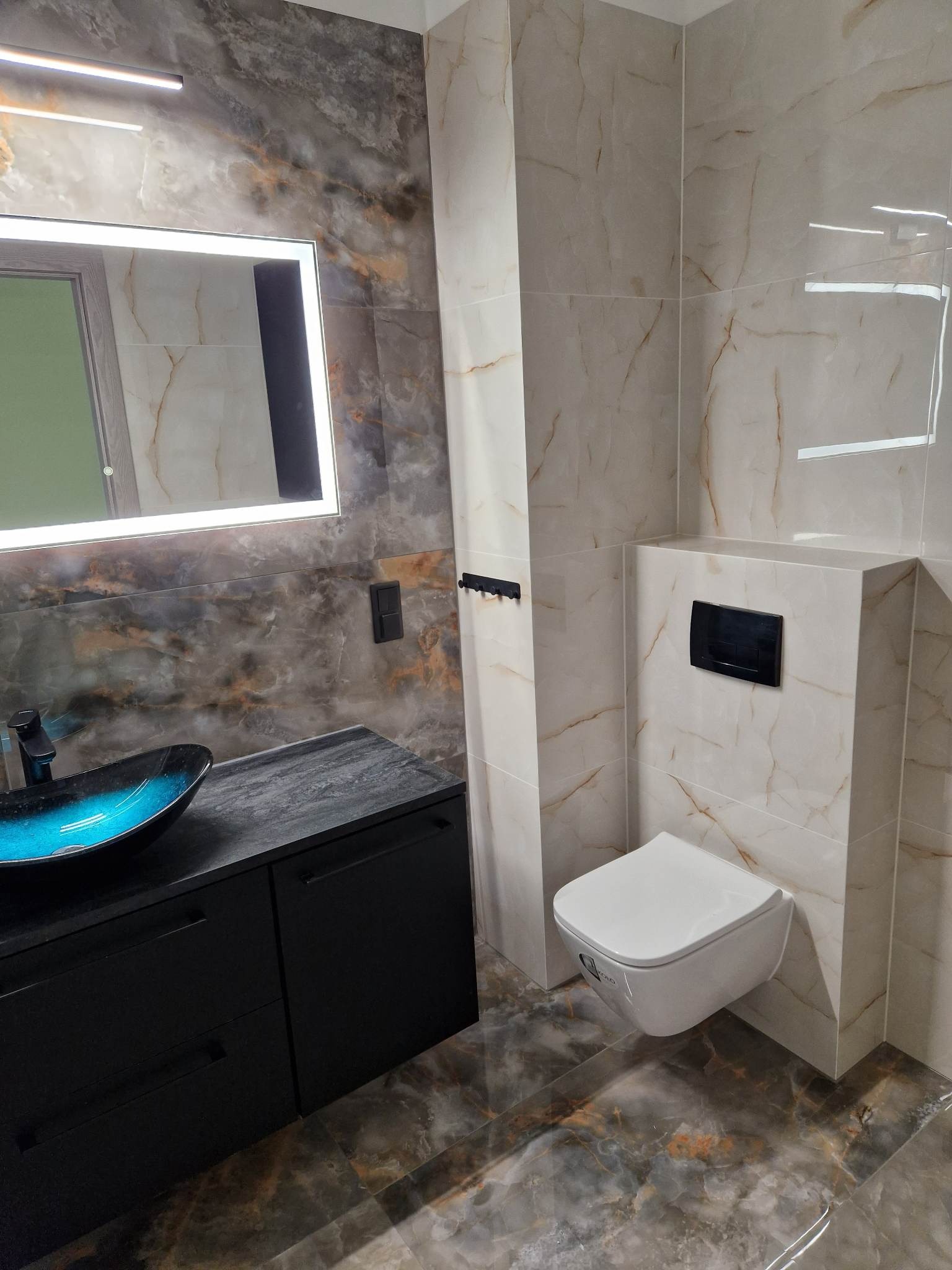 Nowoczesna łazienka z marmurowymi płytkami, podwieszaną toaletą, czarną szafką z umywalką w kolorze turkusowym i podświetlanym lustrem.