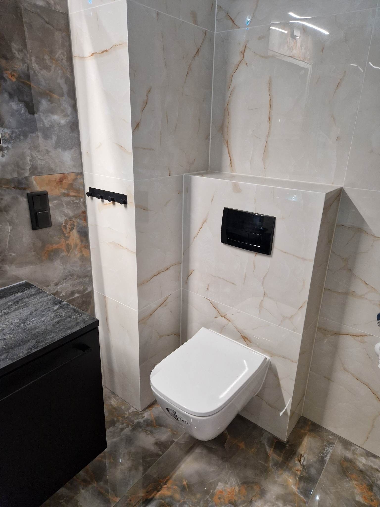 Nowoczesna łazienka z podwieszanym WC, marmurowymi płytkami ściennymi w odcieniach beżu i brązu, czarnym przyciskiem spłukiwania i czarnym wieszakiem na ręczniki, oraz czarną szafką pod umywalką.