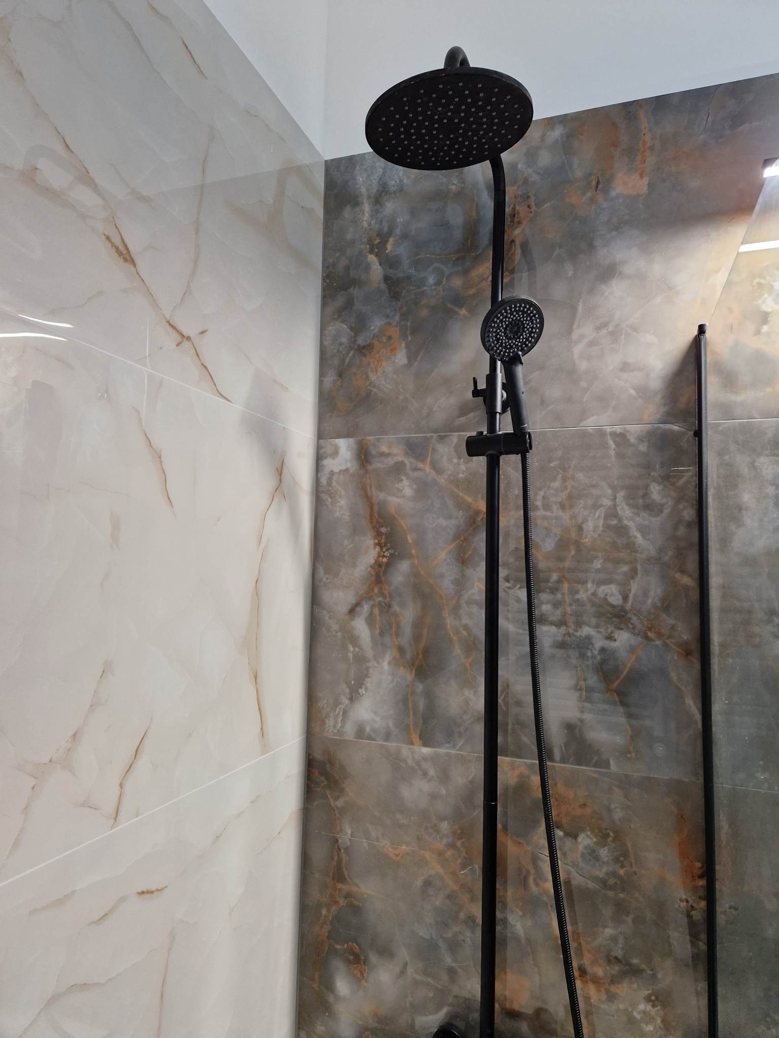 Nowoczesna łazienka z czarną deszczownicą i panelami ściennymi imitującymi marmur w odcieniach szarości i beżu.