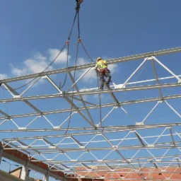 Montaż stalowej konstrukcji dachu hali przez dwóch pracowników w kaskach i kamizelkach odblaskowych, z wykorzystaniem łańcuchów podwieszonych do dźwigu na tle błękitnego nieba.