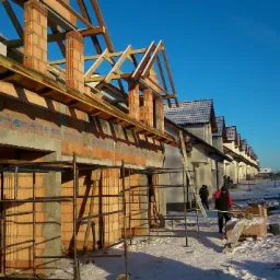 Wznoszenie ścian z czerwonej cegły na budowie szeregu domów w zimowej scenerii, widoczne rusztowania i drewniana konstrukcja dachu.