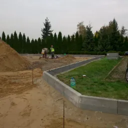 Przygotowanie terenu pod budowę: wyrównany teren z piaskiem, ułożone obrzeża betonowe, pracownicy przy betoniarce, w tle rząd tuj i zaparkowany czarny samochód.