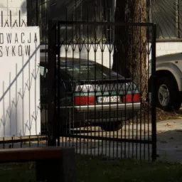 Plakat 'Renowacja Klasyków' obok czarnego auta za metalowym ogrodzeniem i srebrnego SUV-a. Widok z zewnątrz, skupienie na renowacji aut.