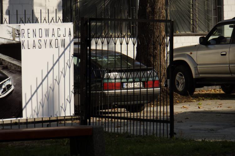 Plakat 'Renowacja Klasyków' obok czarnego auta za metalowym ogrodzeniem i srebrnego SUV-a. Widok z zewnątrz, skupienie na renowacji aut.