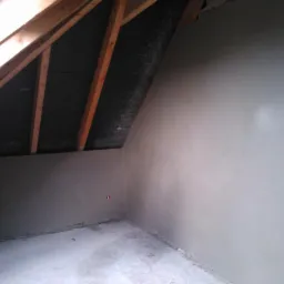 Pokój na poddaszu po nałożeniu gładzi, widoczne drewniane elementy konstrukcji dachu i gładkie ściany w szarym odcieniu. Widoczne gniazdko elektryczne w ścianie.