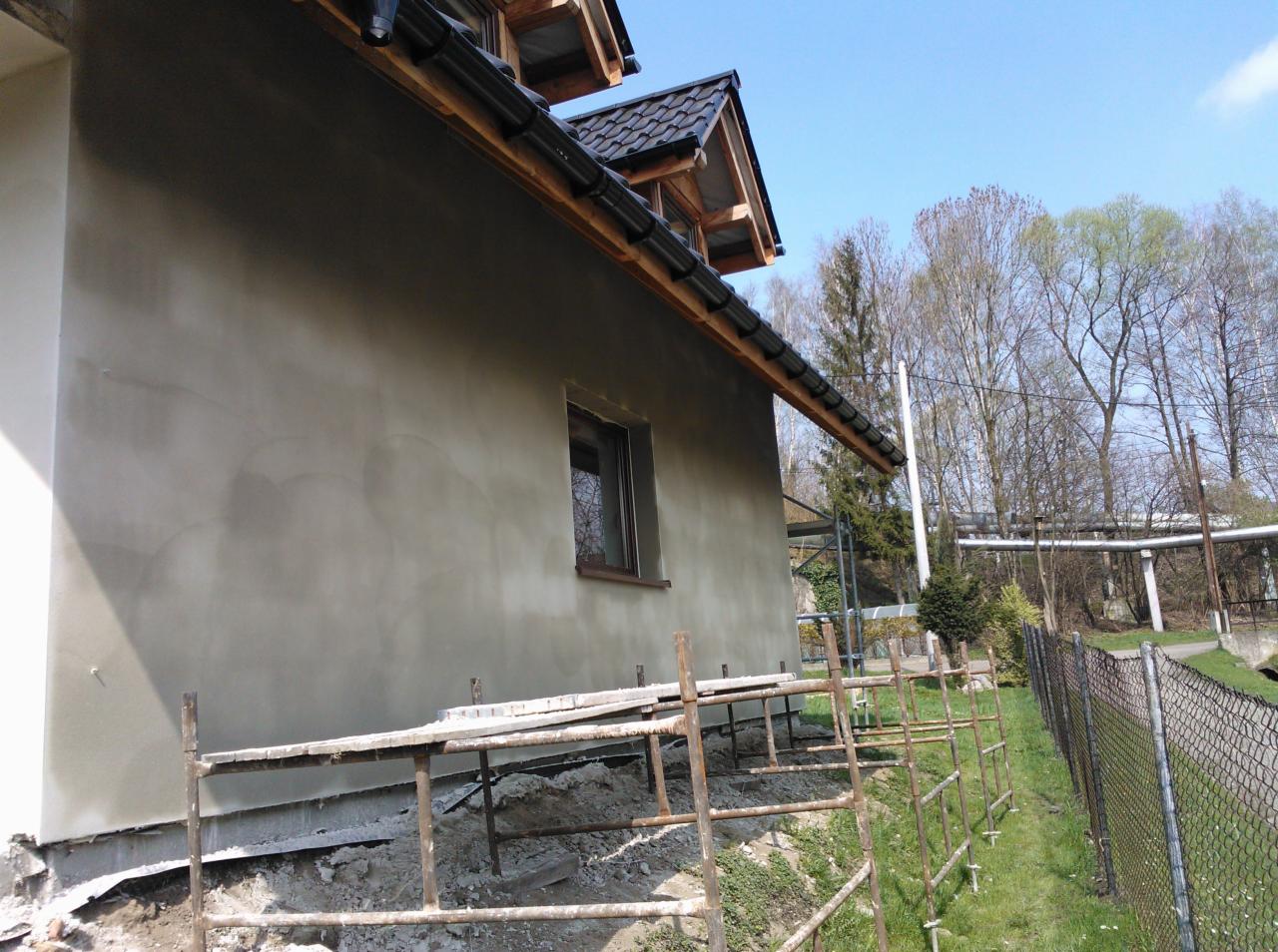 Świeżo otynkowana ściana domu jednorodzinnego z drewnianą konstrukcją dachu, widoczna z rusztowaniami i elementami ogrodu w tle, w słoneczny dzień.