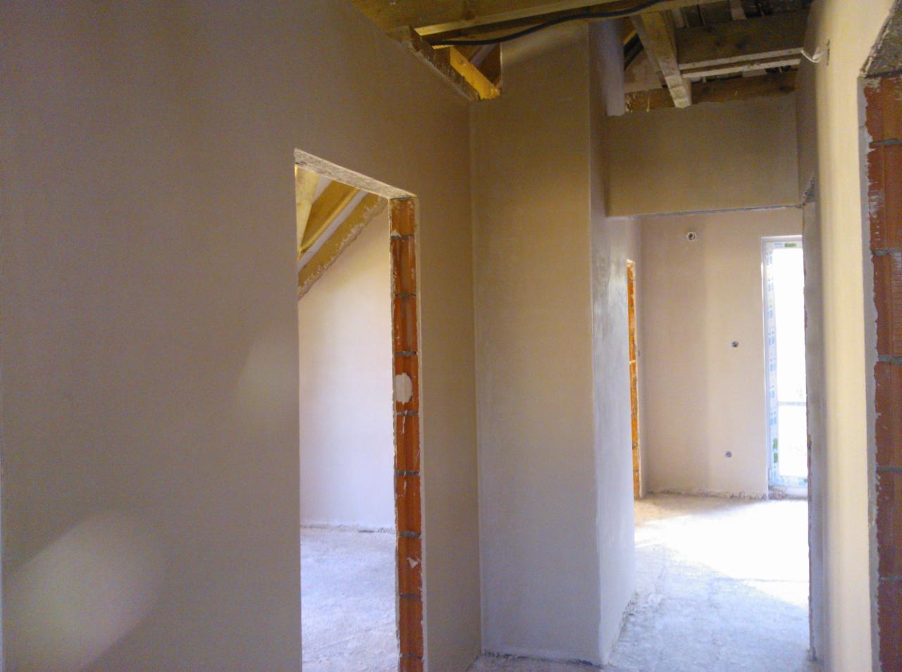 Gładkie, jasne tynki w surowym wnętrzu domu, widoczne fragmenty drewnianej konstrukcji dachu i muru z cegieł. Nowe tynki na ścianach i sufitach.