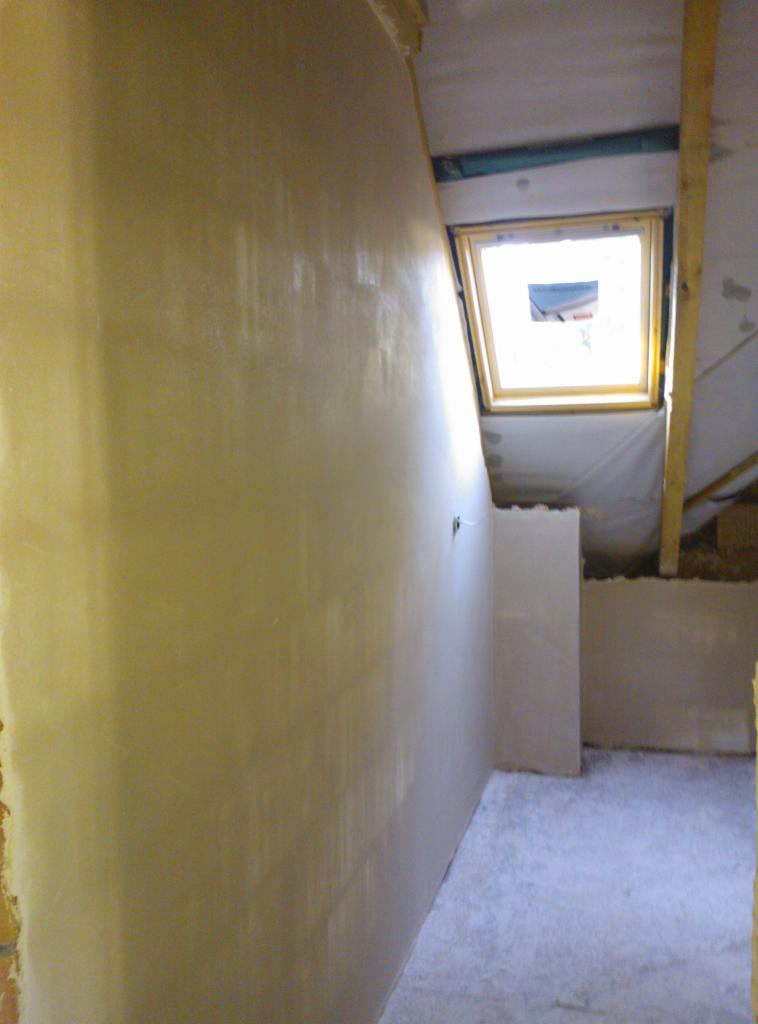 Świeżo nałożony, jasny tynk na ścianie poddasza z oknem dachowym w drewnianej ramie. Widoczne fragmenty konstrukcji dachu i resztki materiałów budowlanych.