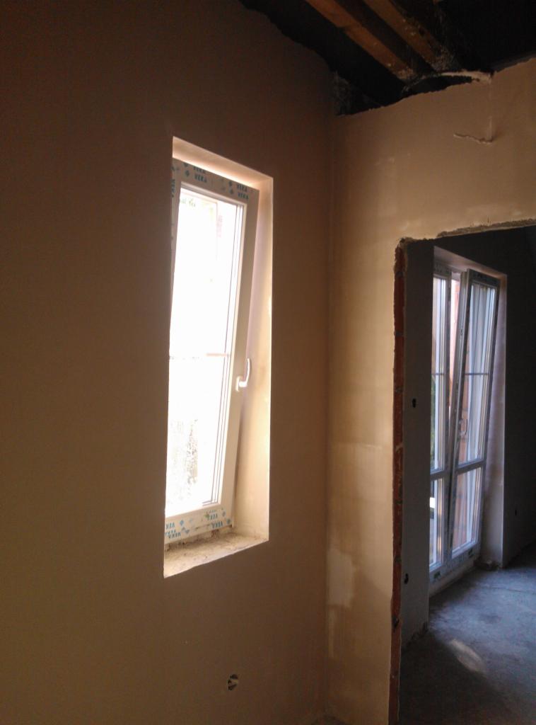 Świeżo otynkowane ściany w jasnym odcieniu beżu, z widocznym oknem i otwartym przejściem do innego pomieszczenia. Widoczne niedoskonałości w górnej części ściany.