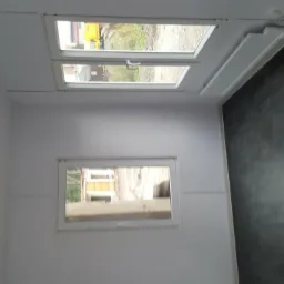 Wnętrze kontenera biurowego z dwoma oknami, grzejnikiem i oświetleniem sufitowym, widok na zewnątrz przez okna.