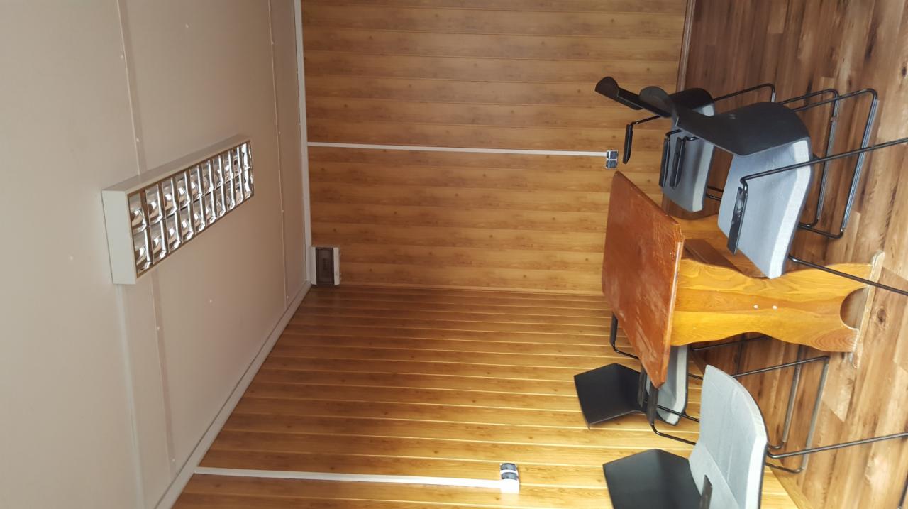 Wnętrze biurowe z drewnianą boazerią i oświetleniem rastrowym, widoczne krzesła biurowe z czarnymi siedziskami i drewniany stół.