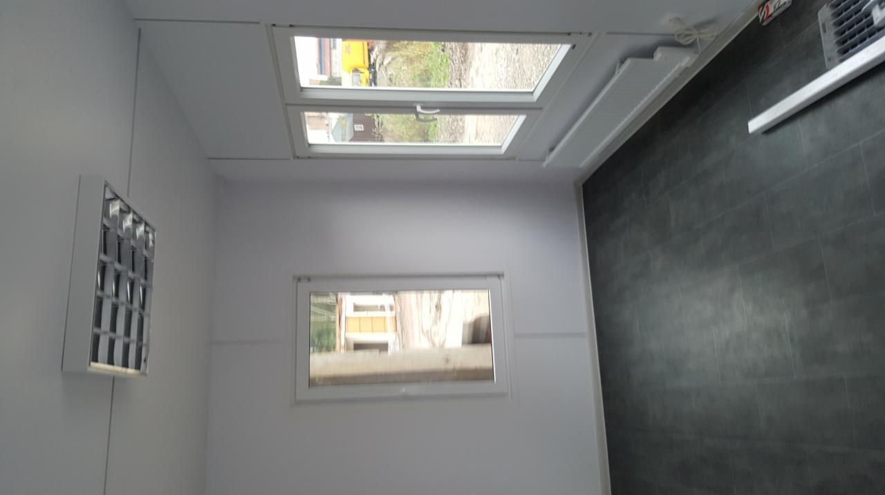 Wnętrze kontenera biurowego z dwoma oknami, grzejnikiem i oświetleniem sufitowym, widok na zewnątrz przez okna.