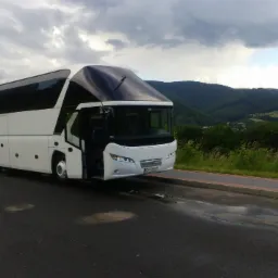 Biały autokar turystyczny z otwartymi drzwiami na tle zielonych wzgórz i pochmurnego nieba.