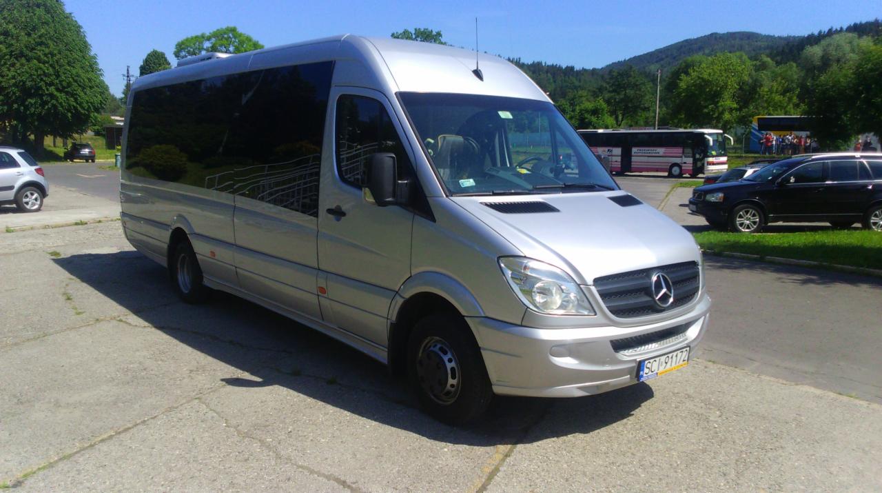 Srebrny bus Mercedes-Benz Sprinter z przyciemnianymi szybami, zaparkowany na betonowym placu w słoneczny dzień, z widocznymi innymi pojazdami i zielenią w tle.
