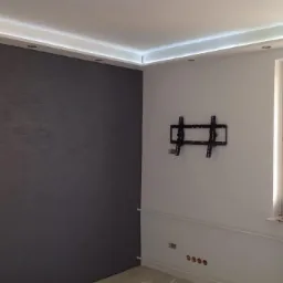 Szara ściana z efektem dekoracyjnym, biała ściana z uchwytem na TV, listwa LED pod sufitem, okno z parapetem i grzejnikiem. Wnętrze po remoncie.