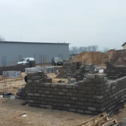 Wznoszenie ścian fundamentowych z bloczków betonowych na placu budowy domu jednorodzinnego. Widoczne zbrojenia i przygotowane materiały.