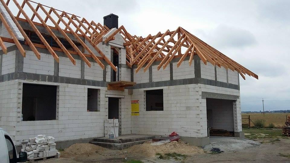 Niedokończony dom z białych bloczków, widoczne drewniane elementy konstrukcji dachu i otwory okienne. Na pierwszym planie materiały budowlane i częściowo widoczny samochód.