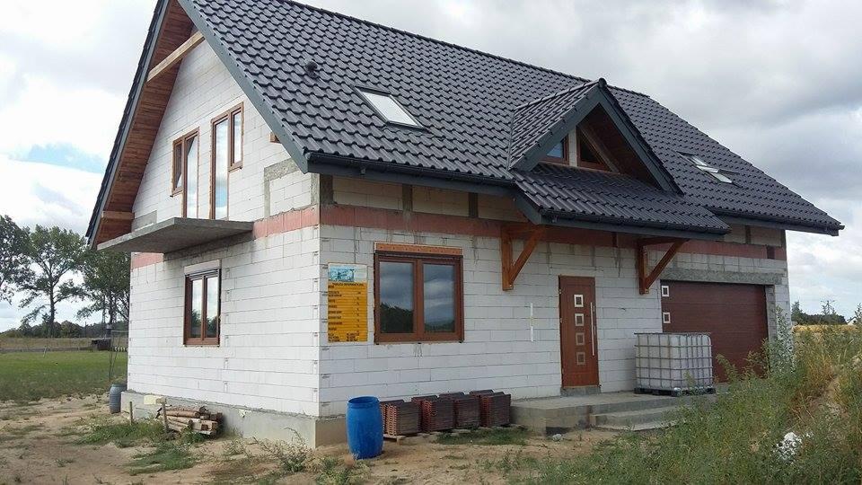 Dwukondygnacyjny dom w trakcie budowy z białego bloczka, z zamontowanymi oknami o brązowych ramach i ciemnym dachem z dachówki. Widoczne elementy konstrukcyjne i materiały budowlane.