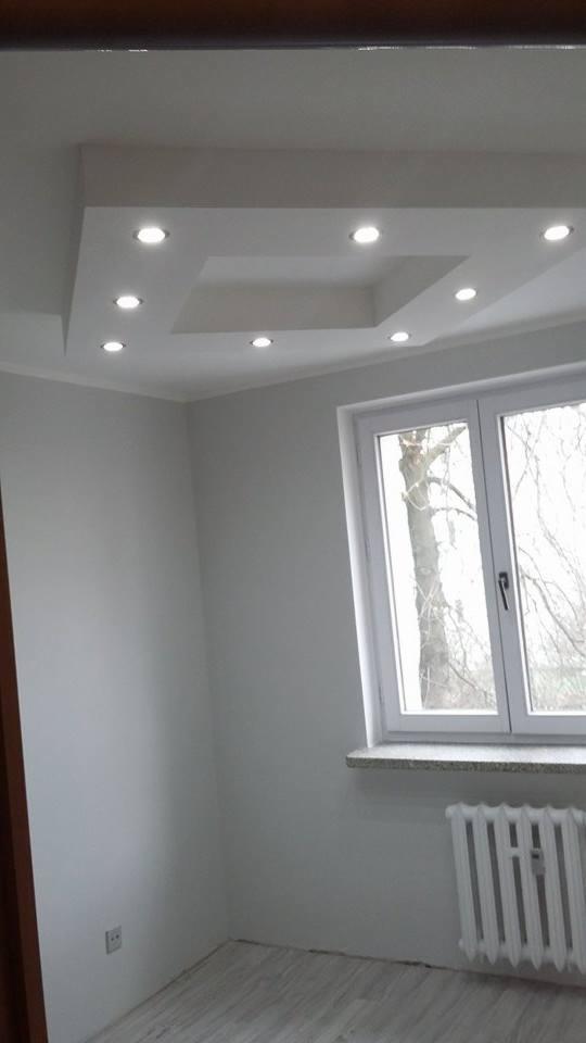 Szary pokój po remoncie: podwieszany sufit z oświetleniem punktowym, okno z parapetem, grzejnik. Ściany pomalowane na szaro, podłoga imitująca drewno.