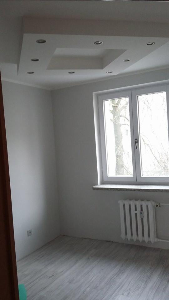 Jasne wnętrze pokoju po remoncie: białe ściany, okno z widokiem na drzewo, panelowa podłoga i grzejnik. Sufit podwieszany z oświetleniem punktowym.