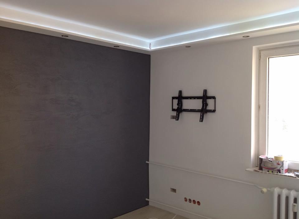 Szara ściana z efektem dekoracyjnym, biała ściana z uchwytem na TV, listwa LED pod sufitem, okno z parapetem i grzejnikiem. Wnętrze po remoncie.