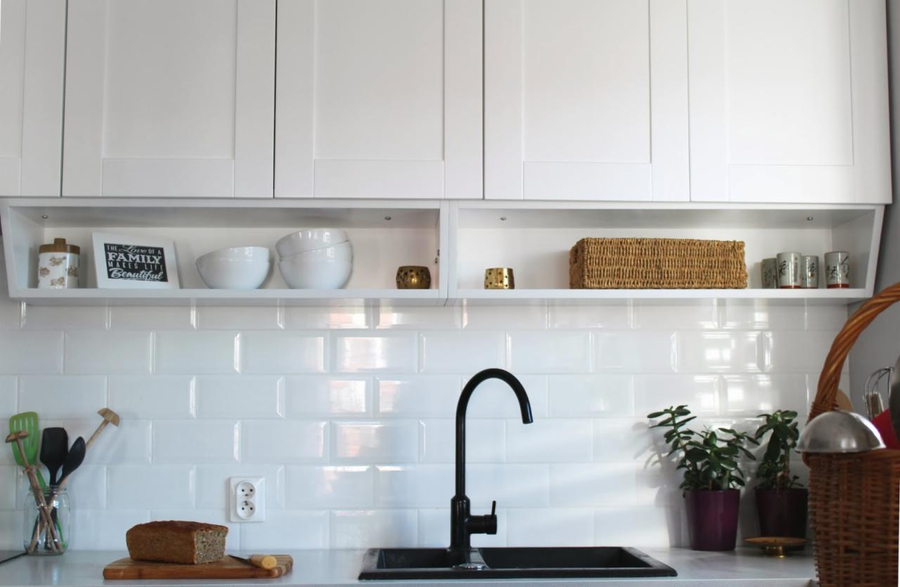 Biała kuchnia z górnymi szafkami, otwartą półką z dekoracjami i czarną armaturą. Ujęcie na płytki ścienne typu metro i minimalistyczny styl.