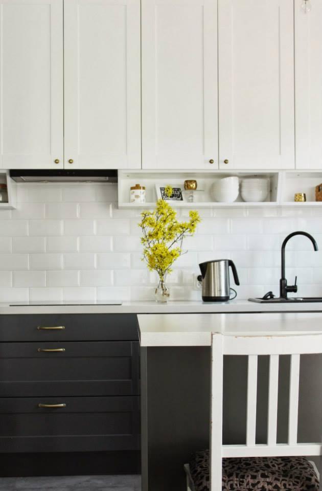 Nowoczesna kuchnia z białymi szafkami górnymi i grafitowymi dolnymi. Na blacie wazon z żółtymi kwiatami, czarny kran i biała ściana z cegiełek.