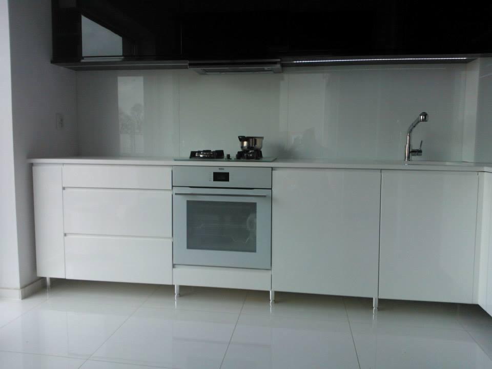 Minimalistyczna kuchnia z białymi, błyszczącymi frontami szafek, jasnym blatem i płytą grzewczą z garnkiem. Nad blatem czarne szafki i oświetlenie LED.