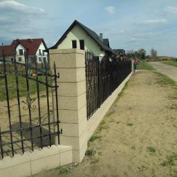 Muka Biuro Zarządzania Nieruchomościami - Eleganckie ogrodzenie kute z elementami dekoracyjnymi, osadzone na słupkach z jasnego kamienia, w tle domy jednorodzinne i polna droga.